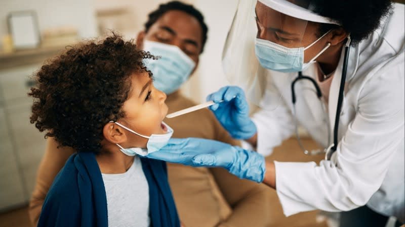 A Black, female physician checks the throat of a pediatric patient while their father looks on.