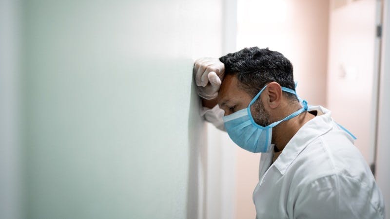 An exhausted physician, wearing a mask and white coat, leans his head against a wall. Recent reports indicate that COVID-19 has exacerbated the already dire circumstances surrounding physician burnout and depression. 